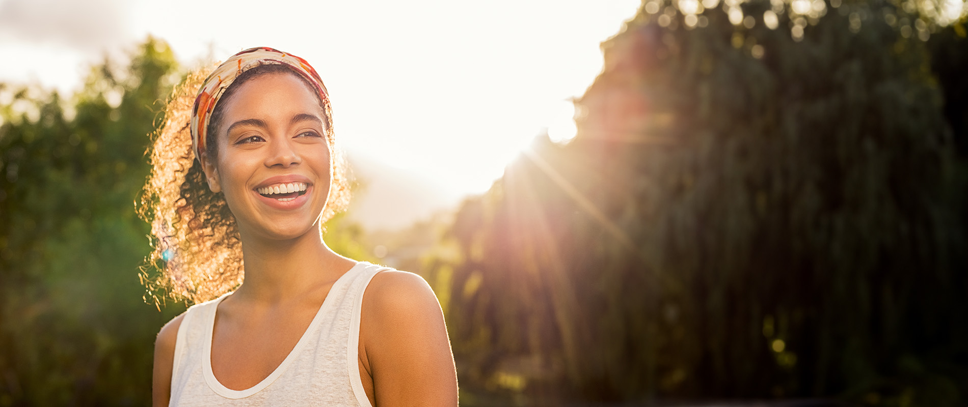 The image shows a person smiling at the camera with sunlight filtering through trees behind them, capturing a moment of joy during daylight.